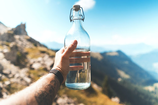 Man holding a glass bottle filled with water against a backdrop of mountains and blue sky.